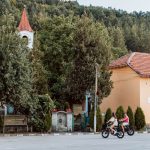 просторна улична сцена с деца каращи колела под дървета и традиционни къщи в България.